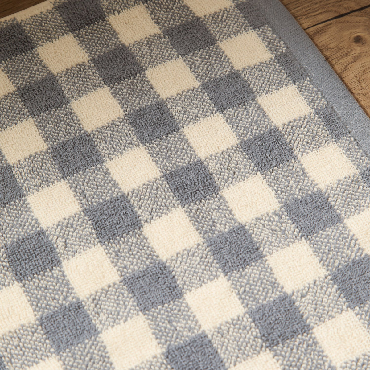 Barton Gingham Cotton Bath Mat in Denim Blue, close-up of the woven denim blue and cream check pattern with looped cotton texture, shown on a wooden floor.