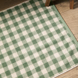 Barton Gingham Cotton Bath Mat in Forest, showing a green and cream check pattern with bound edge, laid on a wooden floor beside furniture legs.