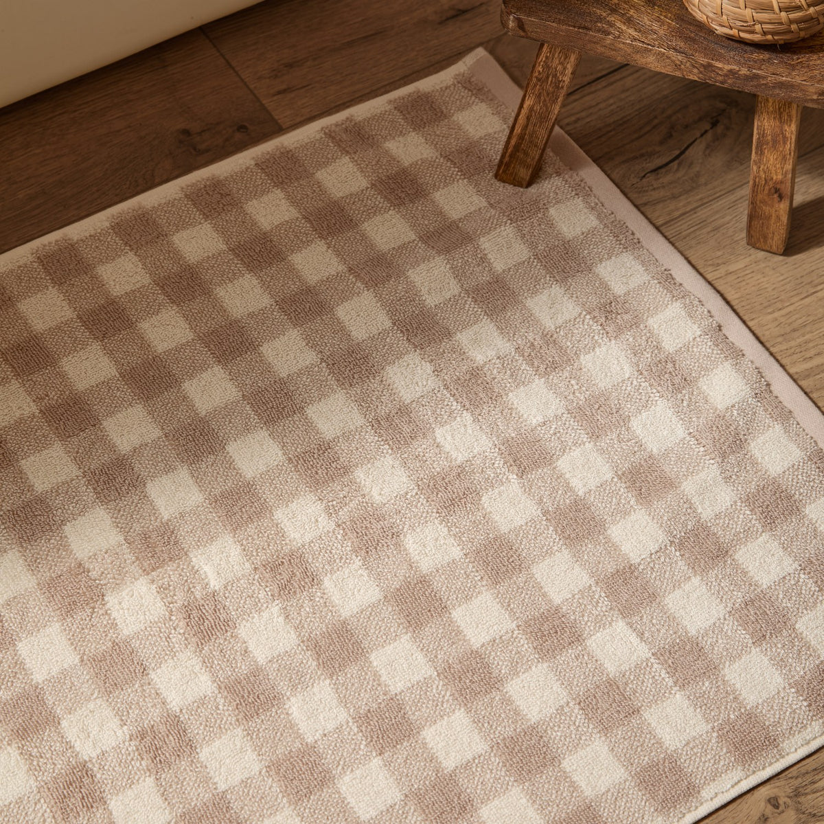 Barton Gingham Cotton Bath Mat in natural taupe and cream check, shown on a wood floor beside a wooden stool.