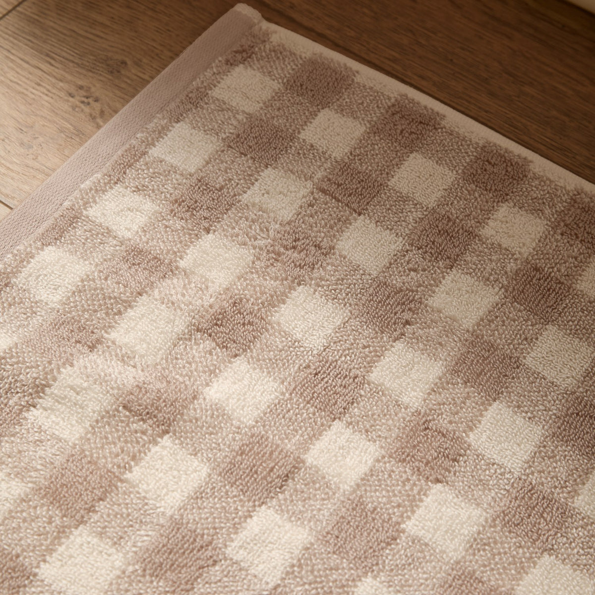 Barton Gingham Cotton Bath Mat in natural, showing woven taupe and cream check pattern with looped cotton pile, corner folded back on a wooden floor.