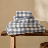 Barton Gingham Cotton Towels in denim blue and cream, folded and stacked in four sizes on a wooden bench in a sunlit room.