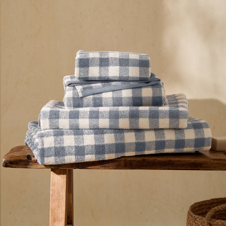 Barton Gingham Cotton Towels in denim blue and cream, folded and stacked in four sizes on a wooden bench in a sunlit room.