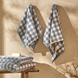 Two Barton gingham cotton towels in denim blue and cream check hang on a wall, with folded towels and soap on a wooden stool below.