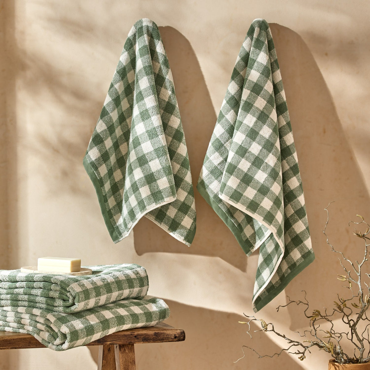 Barton Gingham Cotton Towels in Forest hang on a wall with a matching folded stack and soap on a wooden stool in a bathroom setting.