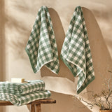 Barton Gingham Cotton Towels in Forest hang on a wall with a matching folded stack and soap on a wooden stool in a bathroom setting.