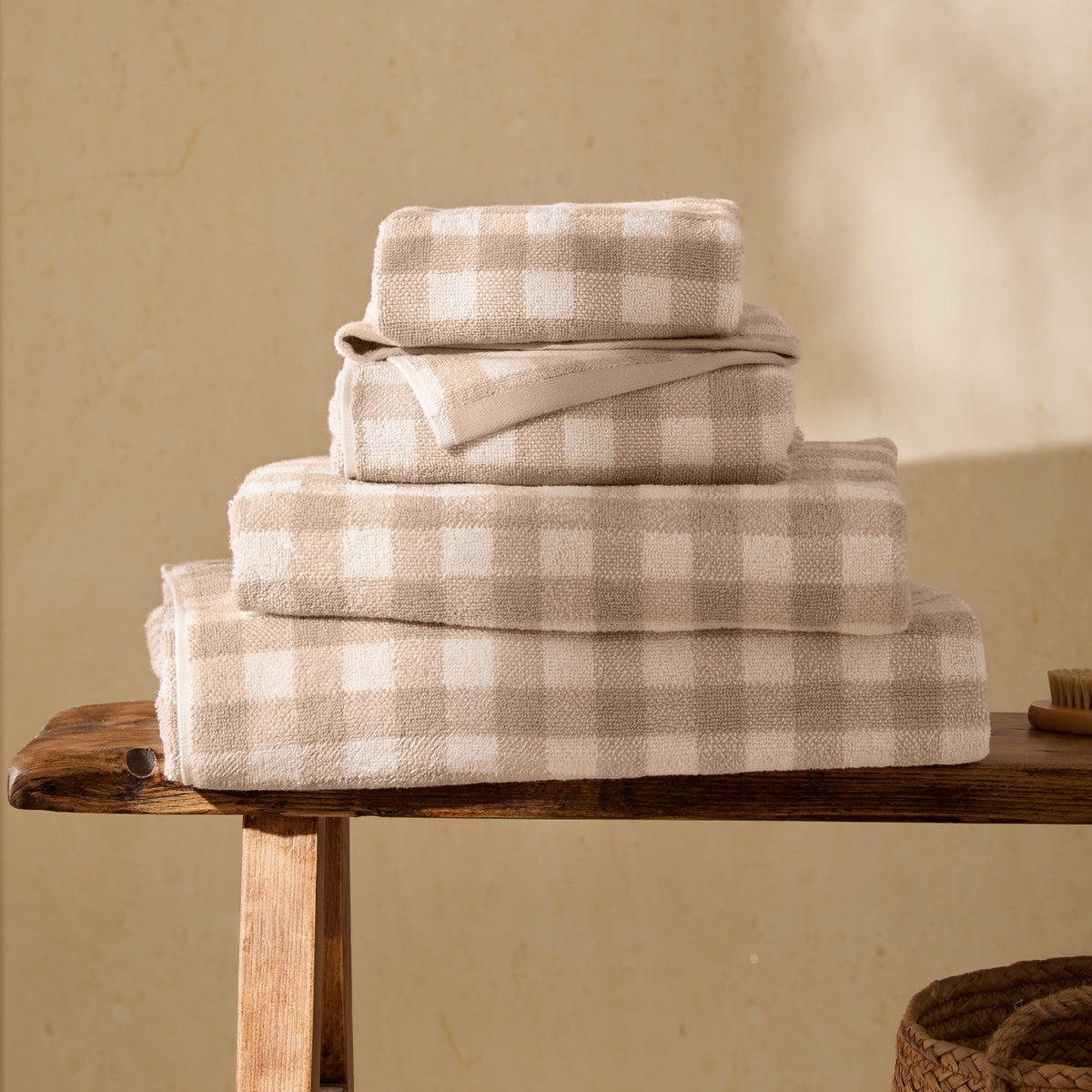 Barton Check Gingham Cotton Towels in natural and cream, stacked in four sizes on a wooden bench, showing the woven check pattern and folded texture.