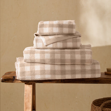 Barton Check Gingham Cotton Towels in natural and cream, stacked in four sizes on a wooden bench, showing the woven check pattern and folded texture.
