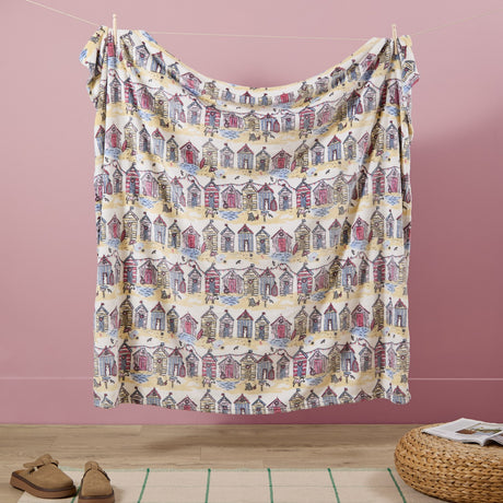 Beach Huts Printed Polyester Throw draped over a washing line against a pink wall, showing a cream fleece with rows of illustrated beach huts in pink, blue and yellow.