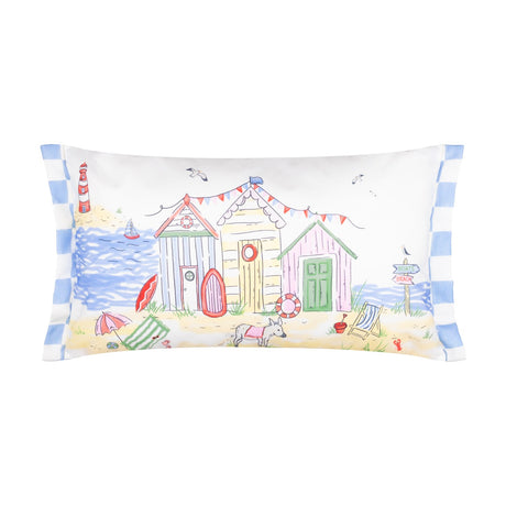 Rectangular polyester cushion with a hand-painted beach scene featuring colourful huts, a lighthouse, sailing boat, bunting and a dog on sand, blue-and-white striped side trim, on a white background.