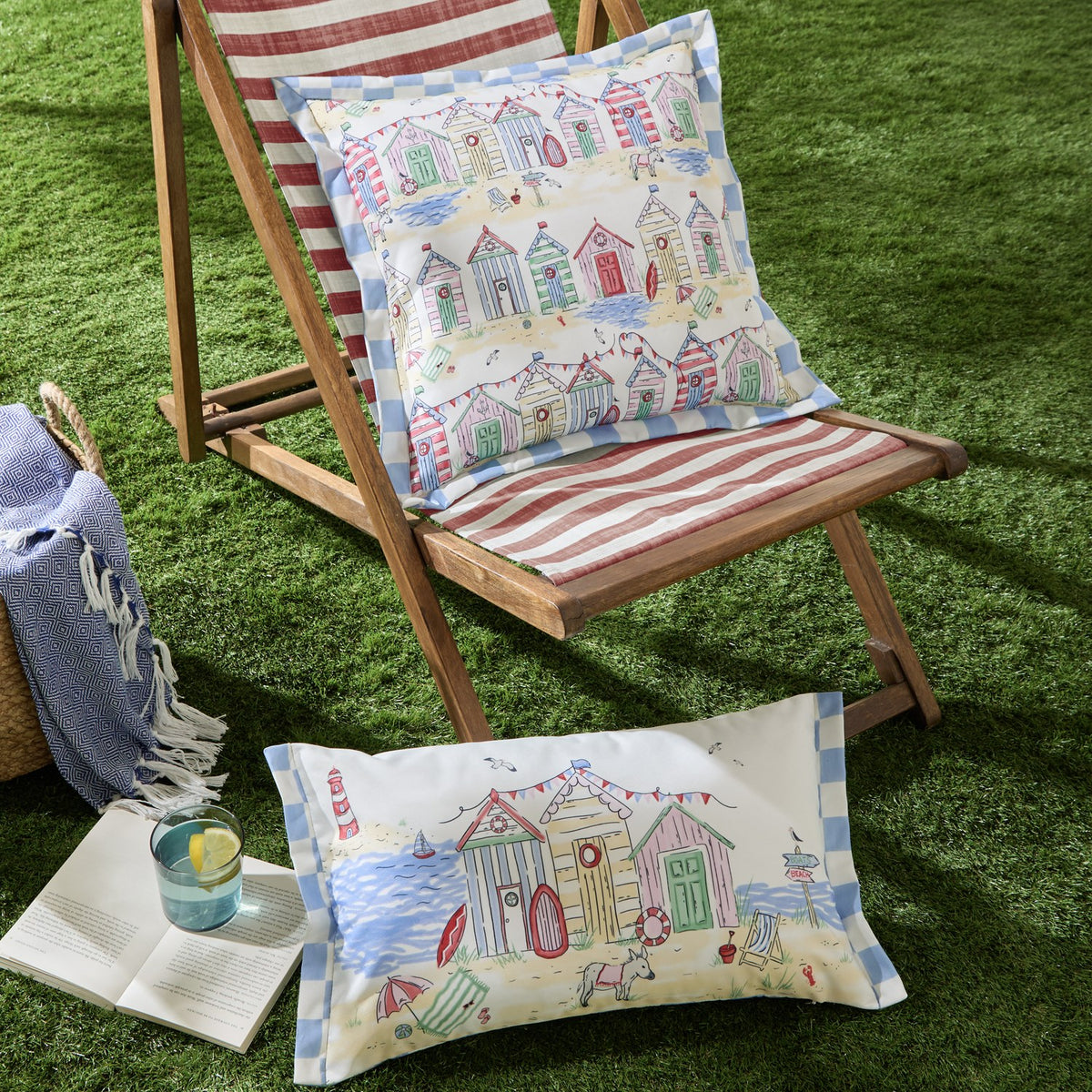 Square and rectangular Beach Huts Gingham polyester cushions with blue check borders, printed with beach huts, bunting and a donkey scene, styled on a striped deckchair on artificial grass.