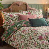 Beatrice Reversible Floral Duvet Cover Set on a bed with matching pillowcases and pink and green cushions, cream and green fabric featuring birds, foliage and berries, against a rattan headboard.
