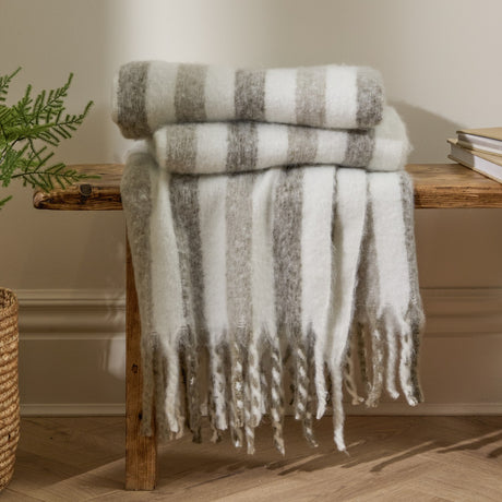 Beechwood Striped Cotton Throw in Heath, cream and grey striped throw with fringed edges, folded and draped over a wooden bench in a living room setting.