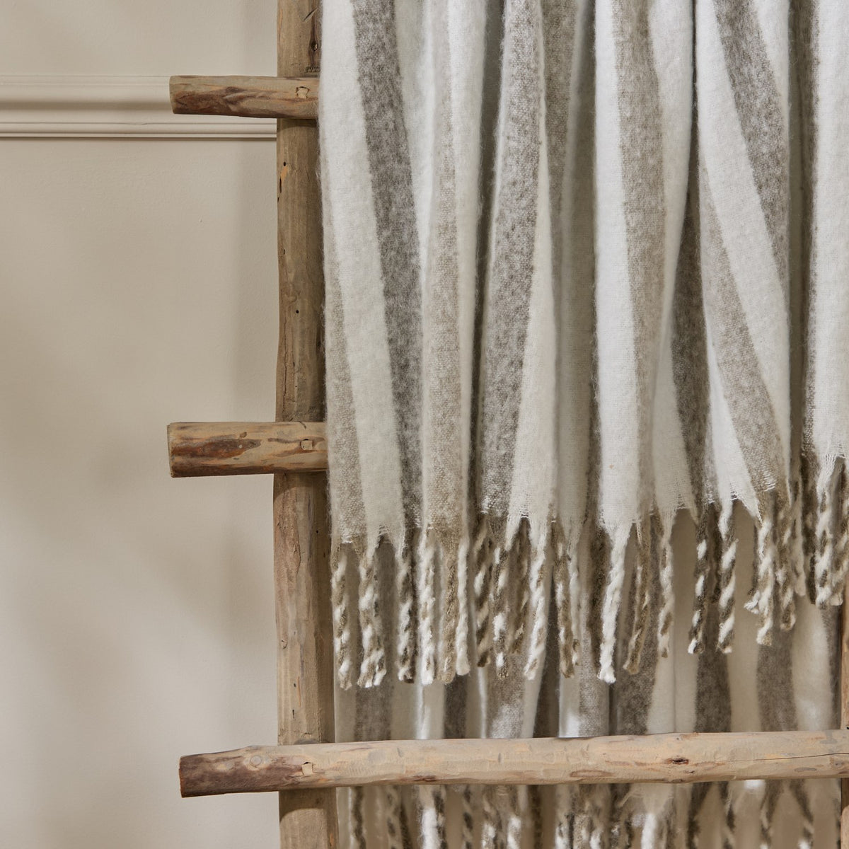 Beechwood Striped Cotton Throw in Heath draped over a wooden ladder rack, showing cream fabric with grey and greige vertical stripes and twisted fringe trim.