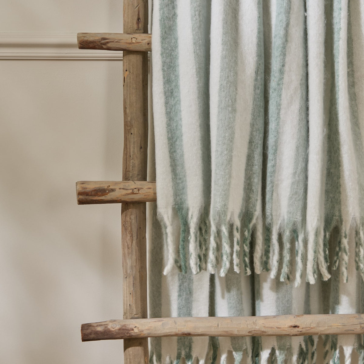 Beechwood Striped Cotton Throw in Moorland Green, brushed cotton with cream and sage vertical stripes and knotted fringe, draped over a wooden ladder.