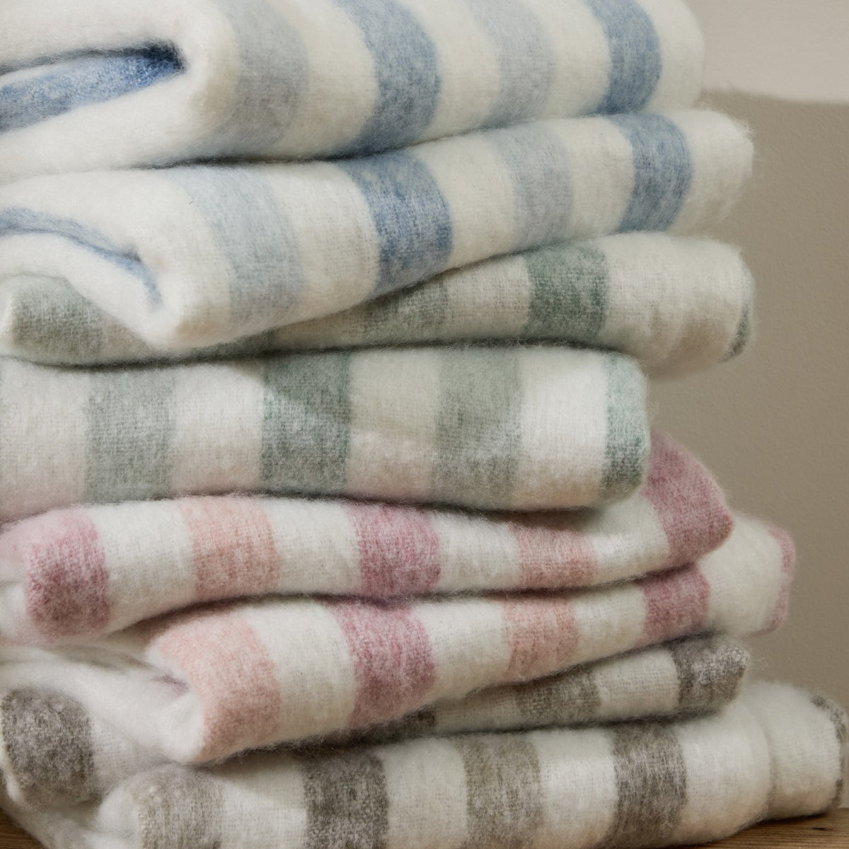 Beechwood Striped Cotton Throw in Moorland Green, folded and stacked with other striped throws in blue, pink, and grey, showing a brushed cotton texture with cream and coloured stripes.