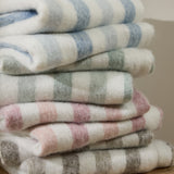 Beechwood Striped Cotton Throw in Moorland Green, folded and stacked with other striped throws in blue, pink, and grey, showing a brushed cotton texture with cream and coloured stripes.