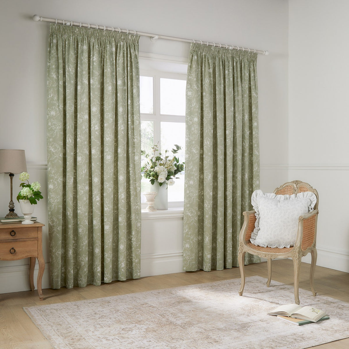 Bella fully lined cotton pencil pleat curtains in sage green with white outline floral pattern, shown framing a window in a living room with a rattan chair and wooden side table.