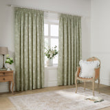 Bella fully lined cotton pencil pleat curtains in sage green with white outline floral pattern, shown framing a window in a living room with a rattan chair and wooden side table.