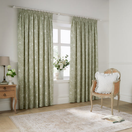 Bella fully lined cotton pencil pleat curtains in sage green with white outline floral pattern, shown framing a window in a living room with a rattan chair and wooden side table.