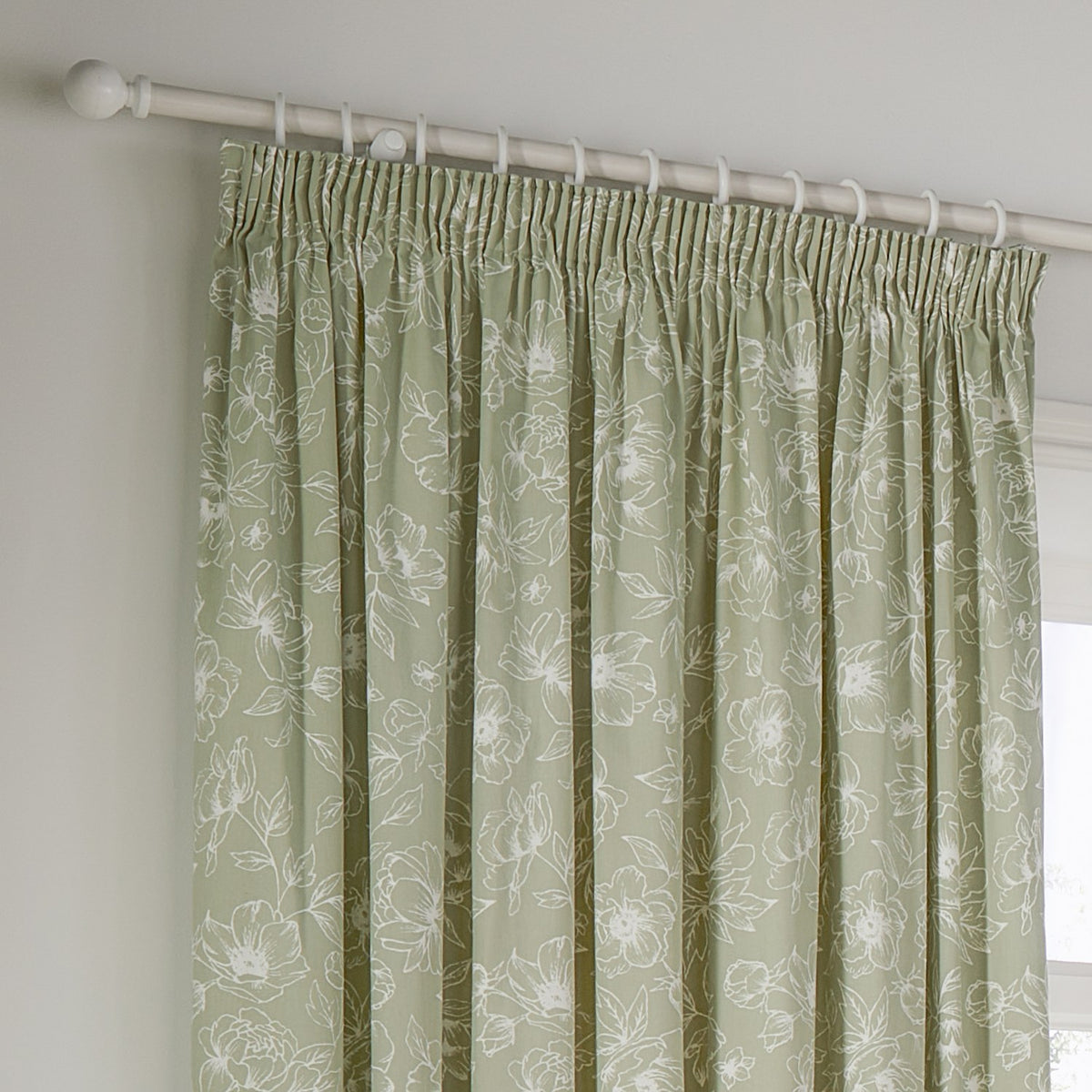 Bella fully lined pencil pleat curtains in sage green cotton with a white outline floral pattern, hung from a white curtain pole near a window.