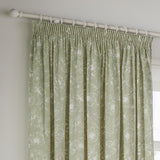 Bella fully lined pencil pleat curtains in sage green cotton with a white outline floral pattern, hung from a white curtain pole near a window.