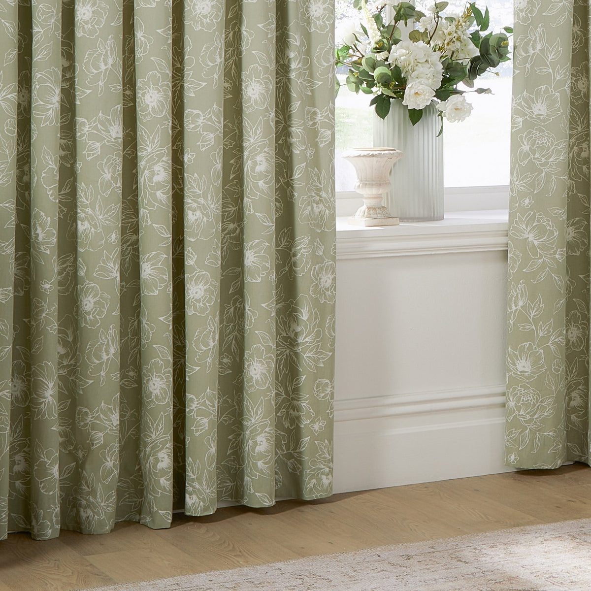 Bella fully lined pencil pleat curtains in sage green cotton with white outline floral pattern, shown pleated beside a window with vase and flowers.