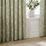 Bella fully lined pencil pleat curtains in sage green cotton with white outline floral pattern, shown pleated beside a window with vase and flowers.
