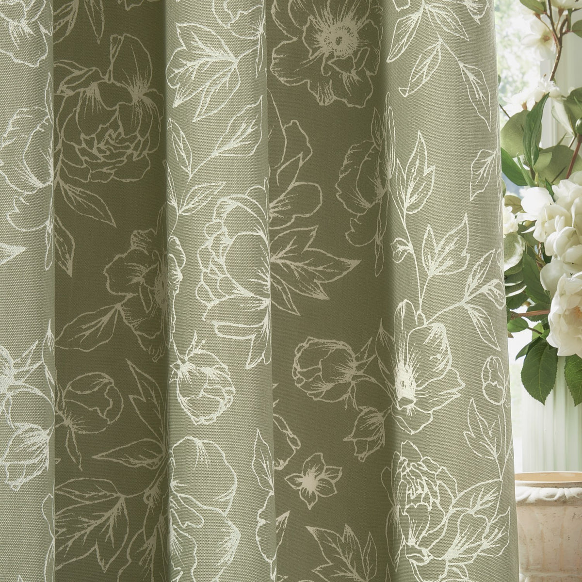 Bella Fully Lined Cotton Pencil Pleat Curtains in sage green, draped in soft folds with a white outline floral peony pattern, shown near a window with white flowers.