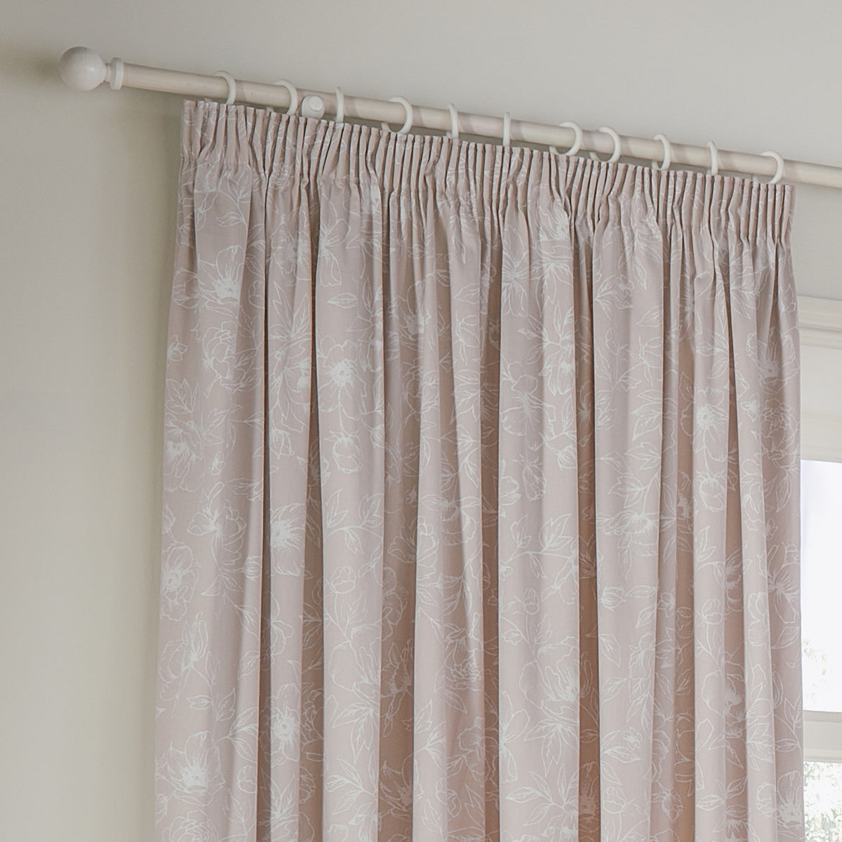 Bella fully lined cotton pencil pleat curtains in natural, showing white floral outline print on a neutral ground, hung from a white pole with curtain hooks near a window.