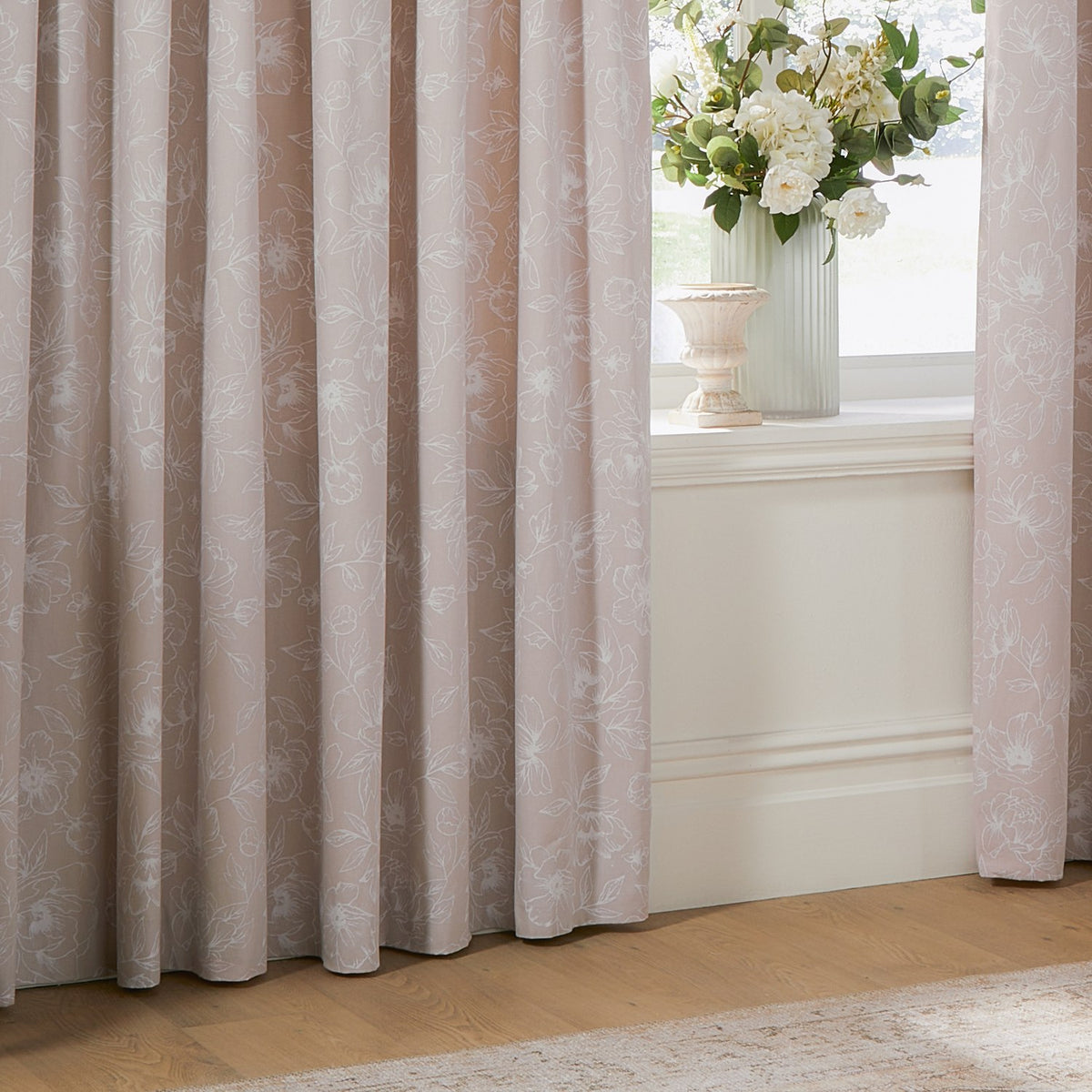 Bella fully lined cotton pencil pleat curtains in natural, showing a white outline floral print on a soft neutral ground, styled beside a window with a vase of flowers.