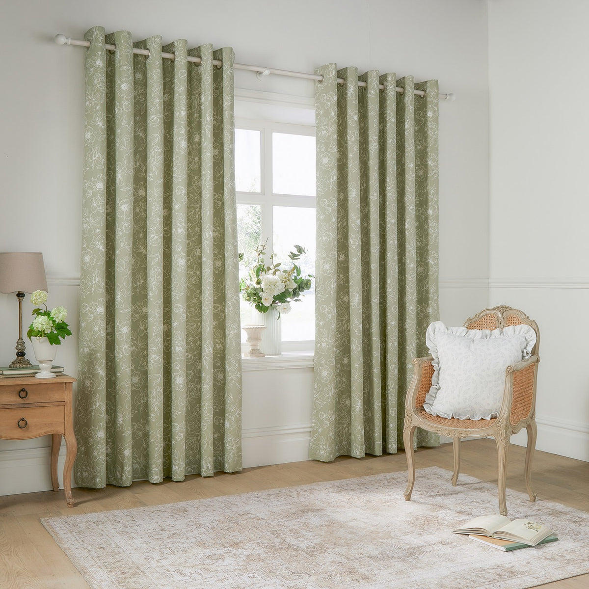 Bella Cotton Fully Lined Eyelet Curtains in sage, showing a pair of floral-print panels on a metal pole in a living room beside a wicker chair and wooden side table.