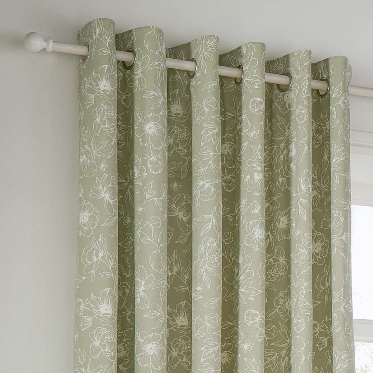 Bella Cotton Fully Lined Eyelet Curtains in Sage, showing eyelet header rings on a pole with white outline floral print on a soft sage cotton ground, hung by a bright window.