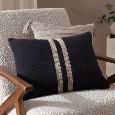 Benton Herringbone Cotton Cushion in black with off-centre pale stripes, shown on a light bouclé armchair alongside a beige tasselled cushion.