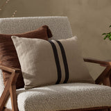 Benton Herringbone Cotton Cushion in natural beige with two dark contrast stripes, shown on a boucle armchair beside a brown textured cushion.