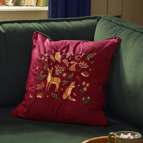 Berry Woods Plain Velvet Cushion in burgundy with embroidered woodland animals, shown on a green sofa in a living room setting.