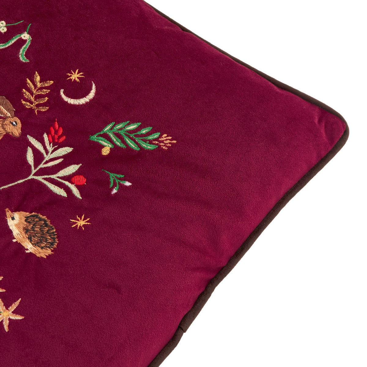 Close-up of the Berry Woods burgundy velvet cushion's corner, showing embroidered hedgehog, hare, foliage, berries and stars with brown piped edge trim.