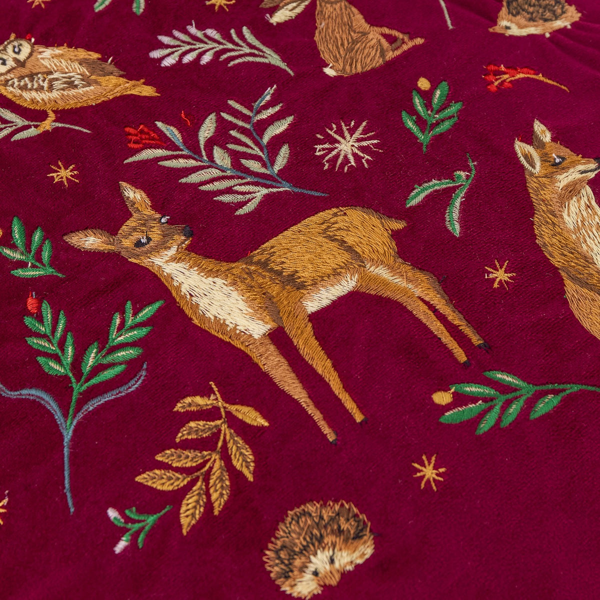 Berry Woods velvet cushion in burgundy, close-up of embroidered fawn, fox, hedgehog and owl among leaves and stars on the matt pile fabric.