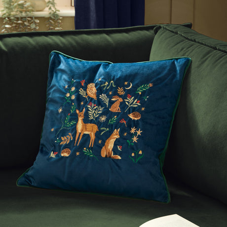 Berry Woods navy velvet cushion with embroidered owl, hare, deer, fox and hedgehogs among leaves and stars, shown on a dark green sofa.