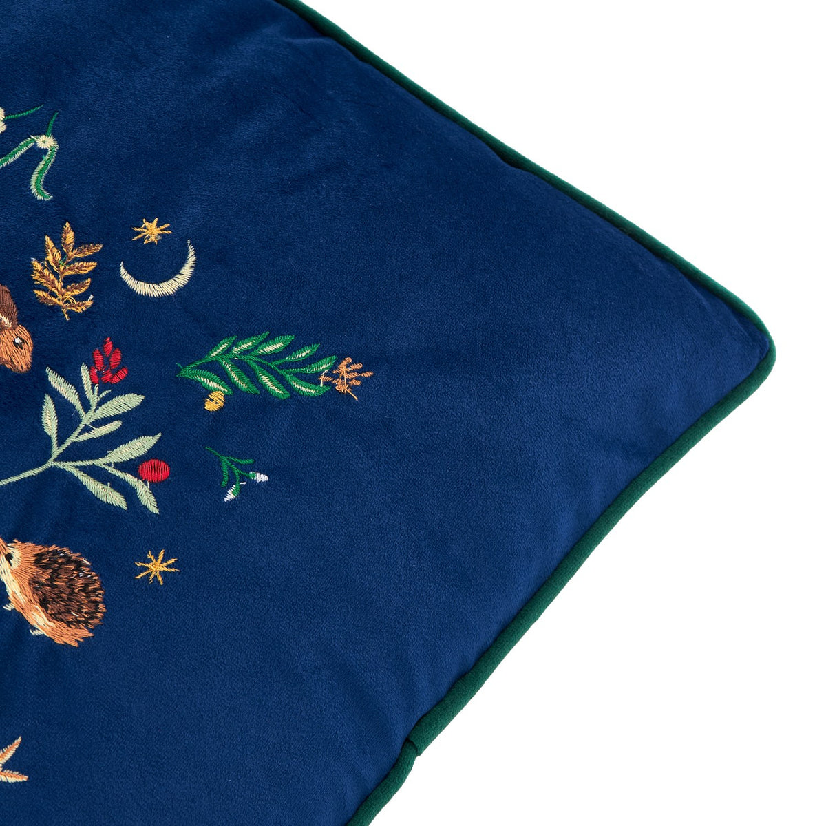 Berry Woods velvet cushion in navy, close-up corner view showing green piped edging and embroidered woodland motifs including a hedgehog, leaves, berries and stars.