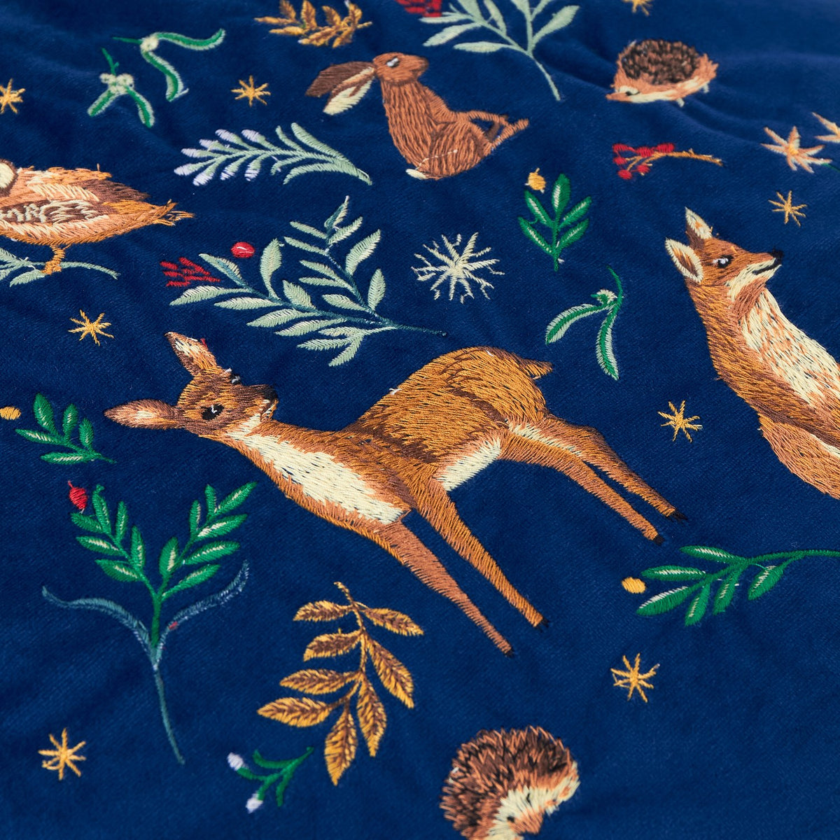 Berry Woods velvet cushion in navy, close-up of embroidered leaping deer, fox, hedgehog and foliage motifs in amber and green thread on the plush fabric.