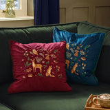 Berry Woods velvet cushion in navy with embroidered owl, hare, deer, fox and hedgehogs, shown on a green sofa beside a matching red cushion.