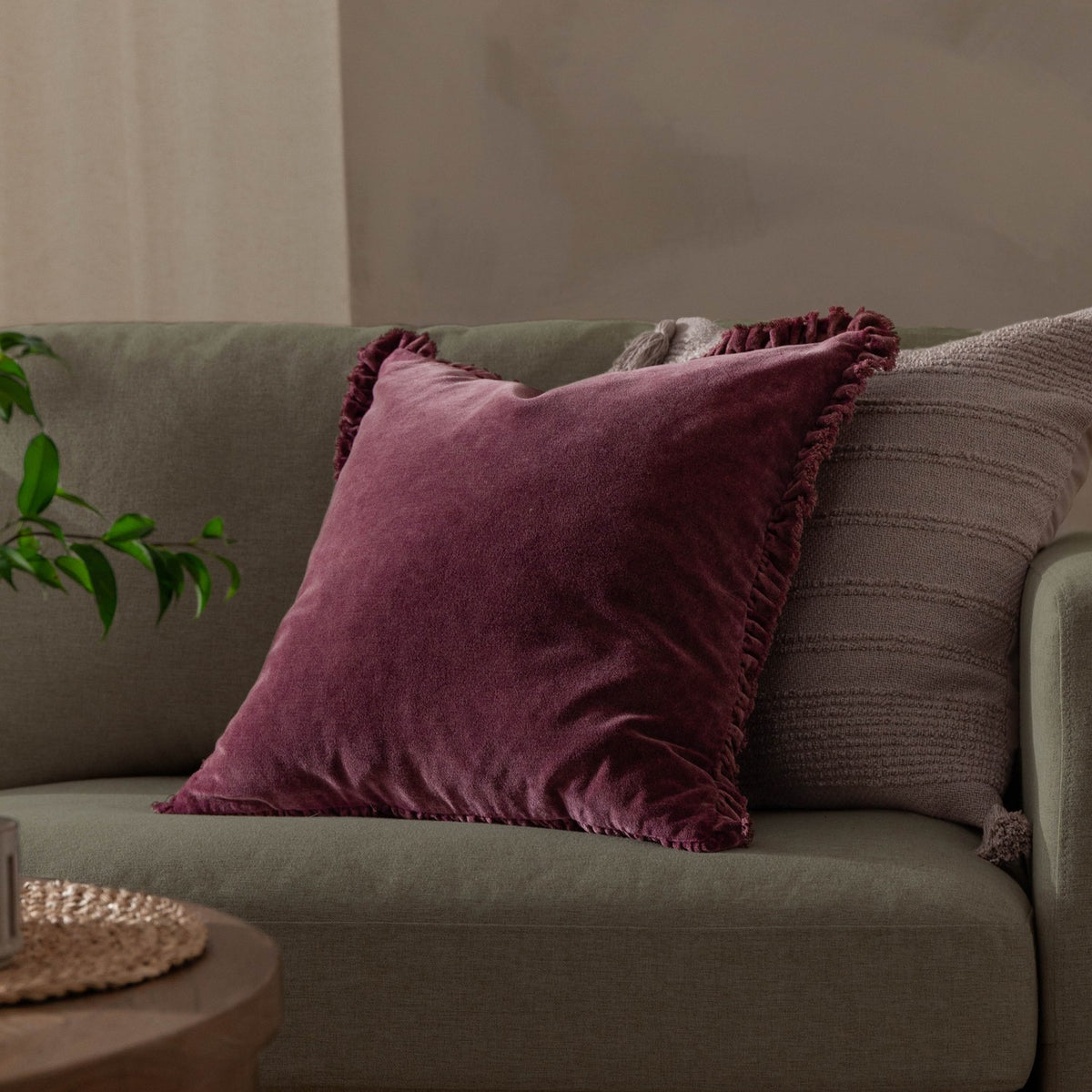 Bertie Velvet Cotton Cushion in plum-burgundy with frayed edge fringe, shown on a sage green sofa alongside a textured grey cushion.