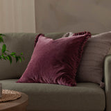 Bertie Velvet Cotton Cushion in plum-burgundy with frayed edge fringe, shown on a sage green sofa alongside a textured grey cushion.