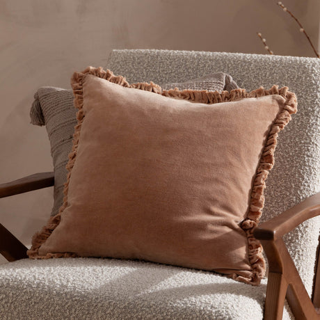 Bertie Velvet Cotton Cushion in peach with ruffled edge trim, shown on a woven armchair beside a grey patterned cushion.