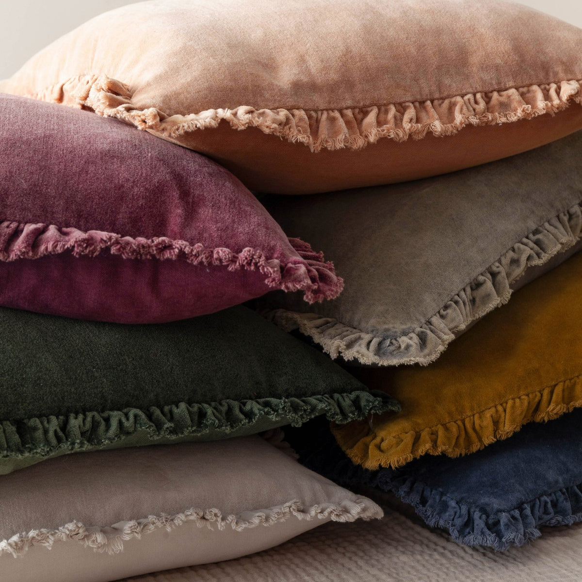 Bertie Velvet Cotton Cushion in peach with frayed-edge ruffle trim, shown stacked with mixed-colour velvet cushions including plum, green, grey, mustard and navy.