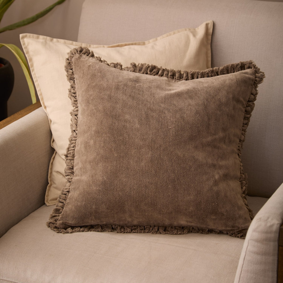 Bertie Velvet Cotton Cushion in chocolate brown with a frayed fringe edge, shown on a beige sofa layered over a cream cushion.