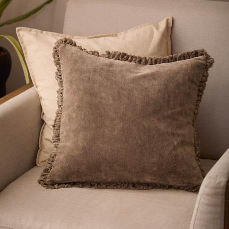 Bertie Velvet Cotton Cushion in chocolate brown with a frayed fringe edge, shown on a beige sofa layered over a cream cushion.