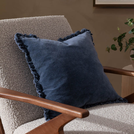 Bertie Velvet Cotton Cushion in mid blue with fringed edge trim, shown on a beige boucle armchair with wooden armrests in a living room setting.