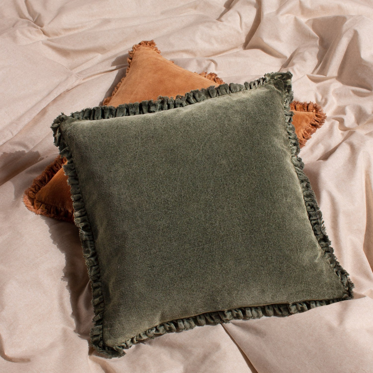 Bertie Velvet Cotton Cushion in moss green with frayed fringed edges, shown on rumpled beige bedding beside a terracotta cushion.