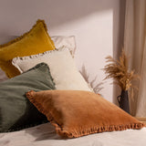 Bertie Velvet Cotton Cushion in moss green with frayed edge trim, styled on a bed among mustard, cream and terracotta cushions with a bedroom backdrop.